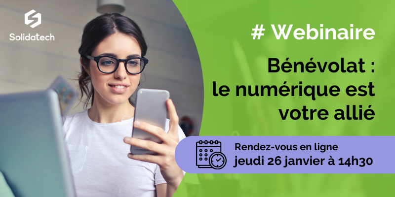 Jeune femme bénévole souriante assise devant un ordinateur et regardant son téléphone portable, qu'elle tient dans sa main.