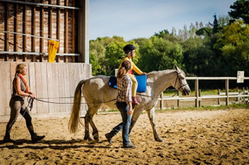 Une enfant en situation de handicap monte à dos de cheval, entourée de deux professionnels de santé