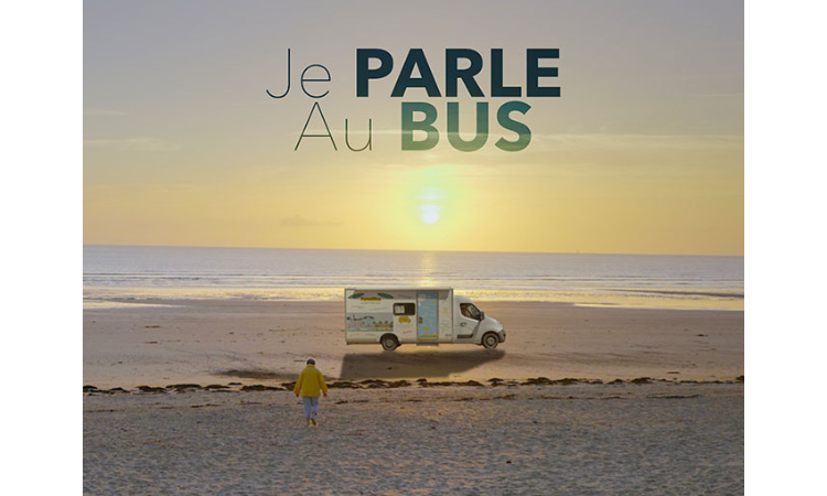 Une personne marche sur une plage au coucher du soleil en direction d’un petit bus stationné sur le sable. En haut de l’image, on peut lire le texte "Je PARLE Au BUS".