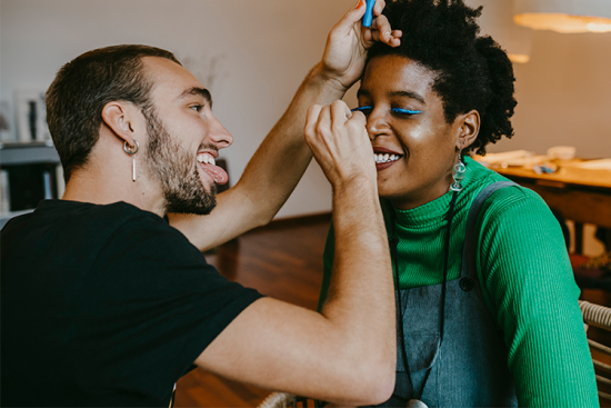 Jeune homme tirant la langue en train de maquiller une jeune femme souriante - © Getty Images