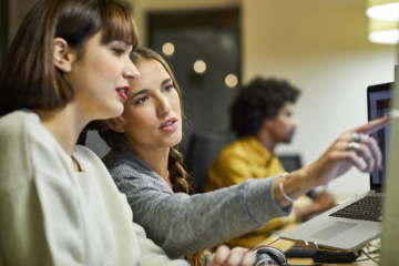 Businesswoman explaining coworker over computer at desk. Female business professionals are discussing in office. They are in smart casuals.